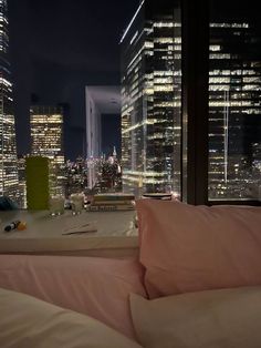 a bed sitting in front of a window next to a night sky line with tall buildings