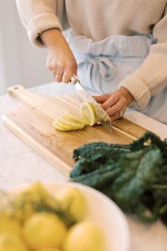 a woman cutting up lemons on top of a wooden cutting board next to greens