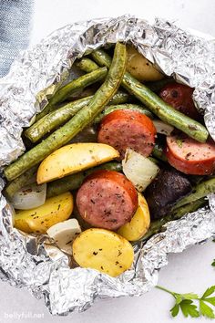 sausage, potatoes and green beans wrapped in tin foil on a white surface with parsley