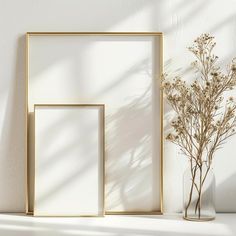 an empty gold frame next to a vase with dried flowers in it on a white wall