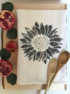 a white towel with a black flower on it next to two spoons and pomegranate