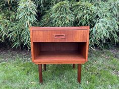 a small wooden table sitting in the grass next to some bushes and trees with one drawer open