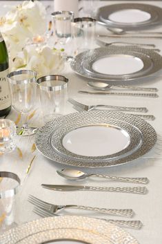 the table is set with silver and white plates, silverware, wine bottle, and flowers