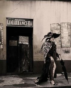 a woman walking down the street in front of a building with an umbrella and feathers on her head