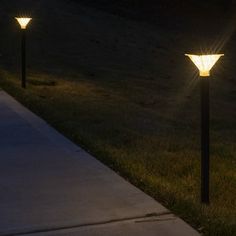 three lights that are on the side of a sidewalk in the grass at night time