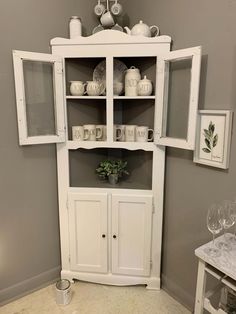 a white china cabinet with glass doors in a corner room filled with wine glasses and other items