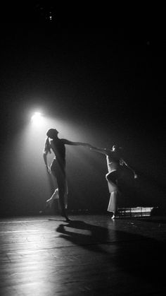 two people are dancing in the dark on stage with lights shining down from behind them