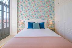 a white bed with blue and pink pillows in a bedroom next to a large window