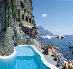 an outdoor swimming pool with chairs and umbrellas next to the cliff side, overlooking the ocean