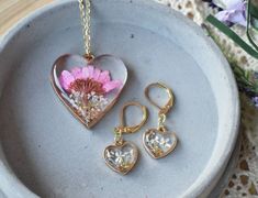 This beautiful handmade botanical jewelry set features a golden heart shaped pendant and earrings, with a purple Daisy and white Queen Anne's Lace flowers, sprouting from a reflective bed of crushed glass. Pressed Flowers that will never die,  preserved forever in crystal clear resin, with a beautiful domed finish.Flower Name & Meaning-  Daisy- new beginnings, rebirth, love. Queen Anne's Lace- safety, beauty, attract loveBezels/ Pendant Frame-  Heart shaped golden frame -1.2" wide x 1.3" tall -A Rose Gold Jewelry Set For Valentine's Day Gift, Valentine's Day Flower Shaped Jewelry Gift, Valentine's Day Flower-shaped Jewelry Gift, Valentine's Day Jewelry With Flower Charm As Gift, Adjustable Flower-shaped Jewelry For Valentine's Day, Flower-shaped Jewelry For Valentine's Day Gift For Her, Adjustable Flower Shaped Jewelry For Valentine's Day, Rose Gold Jewelry Sets For Gifts, Handmade Rose Gold Heart Pendant Jewelry