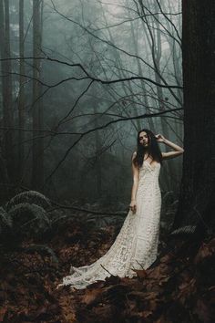 a woman is standing in the woods wearing a white dress and holding her hair back