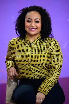 a smiling woman sitting on top of a chair in front of a purple wall and wearing black pants