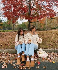 two women sitting on a bench in front of a tree with autumn leaves around them