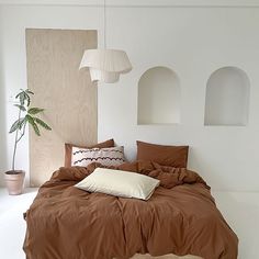 a bed with brown sheets and pillows in a white room next to a potted plant