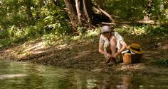 a woman sitting on the bank of a river with a basket and hat in her hand