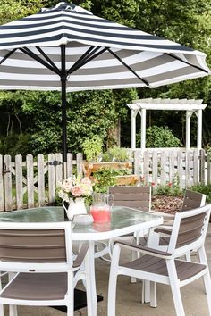 a table with chairs and an umbrella on top of it in front of a white picket fence