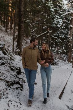 a man and woman are walking through the snow in front of some trees, holding hands