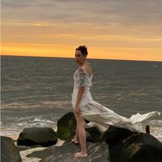 a woman is standing on some rocks by the ocean at sunset with her dress blowing in the wind