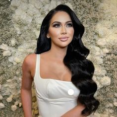 a woman with long black hair wearing a white dress and posing in front of flowers