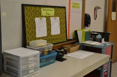 a desk with several file folders on it in an empty classroom area, next to a bulletin board