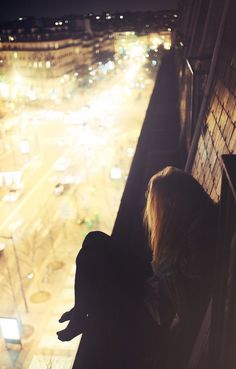 a woman sitting on top of a building next to a window with a dog looking at the street below