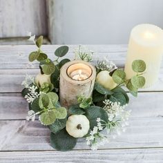 an arrangement of greenery and candles is displayed on a wooden table in front of a white candle