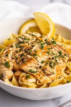 a white bowl filled with pasta and chicken on top of a table next to lemon wedges