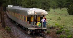 two people standing on the side of a train as it goes down the tracks in the woods