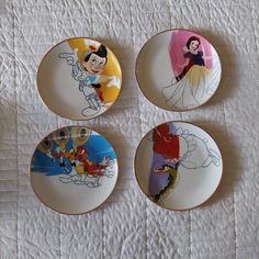 four plates with disney characters painted on them sitting on a white tablecloth covered surface