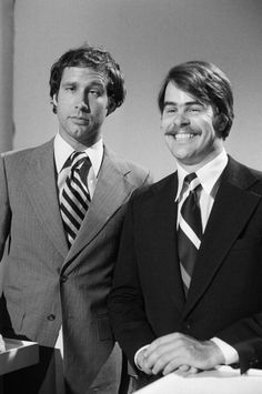 two men in suits and ties standing next to each other with one man smiling at the camera