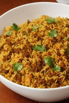 a white bowl filled with yellow rice and meat covered in cilantro leaves on top of a wooden table