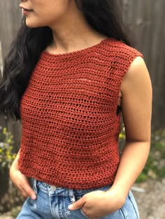 a woman wearing a red crochet top with her hands in her pockets, standing next to a wooden fence