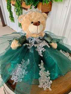 a teddy bear dressed in a green dress sitting on top of a wooden table with greenery