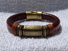 "A lovely combination of an approximate 1\" hammered slider flanked by thinner spacers and rubber rings to hold everything in place.  All on a thick (about 1/4\") band of Regaliz leather and finished with an easy on/easy off magnetic clasp.  The leather featured is a tobacco brown color. The rubber rings are rust-colored. The sliders and clasps are brass.     All clasps and sliders are made from zinc that is mined in Germany or Belgium.  They are certified lead and nickel free. Please select you Brown Leather Bracelets With Brass Hardware, Adjustable Leather Bracelet With Magnetic Closure, Adjustable Brown Leather Bracelet With Brass Hardware, Adjustable Gold Leather Bracelet With Brass Hardware, Adjustable Brown Brass Jewelry, Casual Brown Metal Bracelets, Casual Brown Metal Bracelet, Adjustable Brown Metal Bracelet, Adjustable Brown Metal Bracelets