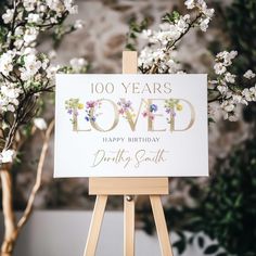 a sign that says 100 years loved sitting on top of a easel next to some flowers