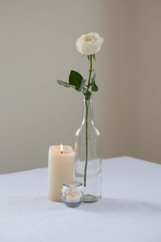 a single white rose in a glass vase next to a lit candle on a table