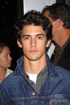 a young man in a denim jacket standing next to other people and looking at the camera