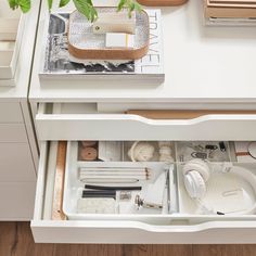 an open drawer with various items in it on top of a white table next to a potted plant