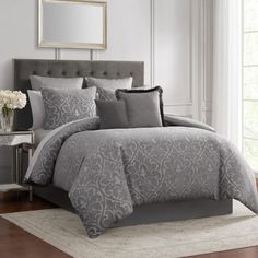 a bed with grey comforter and pillows in a room next to a window,