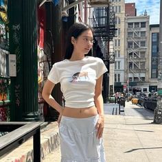 a woman standing on the sidewalk wearing a white skirt and cropped t - shirt