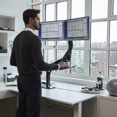 a man standing in front of two computer monitors on top of a desk next to a window