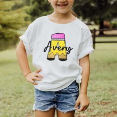 Young girl wearing a white personalized monogram school pencil back to school shirt with a large A that looks like a school pencil with a personalized name written in black script font across the middle of the pencil initial. First Day Of School Shirts Kids, Personalized Short Sleeve T-shirt For Back To School, Customizable T-shirt For Back To School Events, School Spirit Tops With Name Print For School Events, Customizable T-shirt For School Events And Back To School, Customizable School Spirit Tops For School, Back To School Spirit T-shirt With Name Print, Customizable T-shirt For School Events, Customizable School Spirit Tops