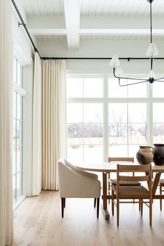 a dining room table and chairs in front of large windows with white drapes on them