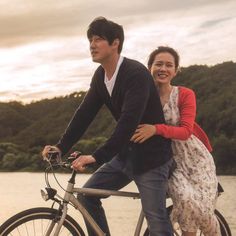 a man and woman riding on the back of a bike next to a body of water