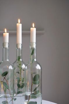 three glass bottles with plants in them sitting on a table next to two lit candles