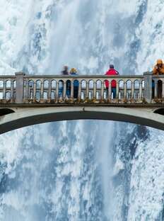 people are standing on a bridge over the water in front of a waterfall, looking up into the sky