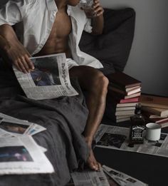 a shirtless man sitting on a bed drinking from a cup and reading the paper