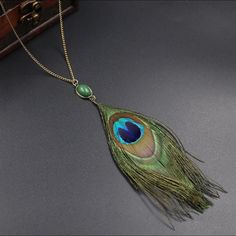 a peacock feather pendant is hanging from a chain on a gray surface with a wooden box in the background