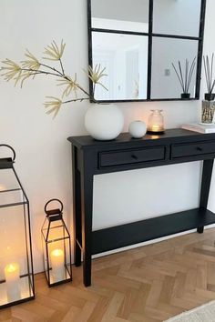 A minimalist Scandinavian-style hallway featuring a sleek black console table with two drawers. The table is decorated with simple white ceramic vases, a glass candle holder, and reed diffusers, exuding a clean, modern vibe. Above the table, a black-framed rectangular mirror reflects light into the space. On the floor, two black-framed glass lanterns with glowing pillar candles add warmth to the neutral palette.