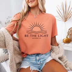 Spread positivity and faith with our "Be the Light" graphic tee! Featuring a beautiful sun design and the powerful scripture from Matthew 5:14, this Christian shirt serves as a daily reminder to shine your light wherever you go. This soft, comfortable t-shirt is perfect for casual wear, faith-based events, church gatherings, or as an inspirational gift for friends and loved ones. Made from high-quality fabric, this Bible verse tee pairs effortlessly with jeans, shorts, or a cozy cardigan. It's a Church Shirt Designs, Bible Verse Tees, Church Shirt, Matthew 5, Bible Verse Shirt, Sun Design, Be The Light, Spread Positivity, Sun Designs